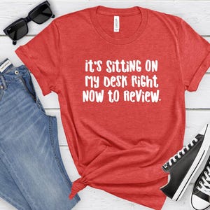 May include: Red t-shirt with the text "It's Sitting On My Desk Right Now To Review" in white. The shirt is paired with blue jeans, black sunglasses, and black Converse sneakers.