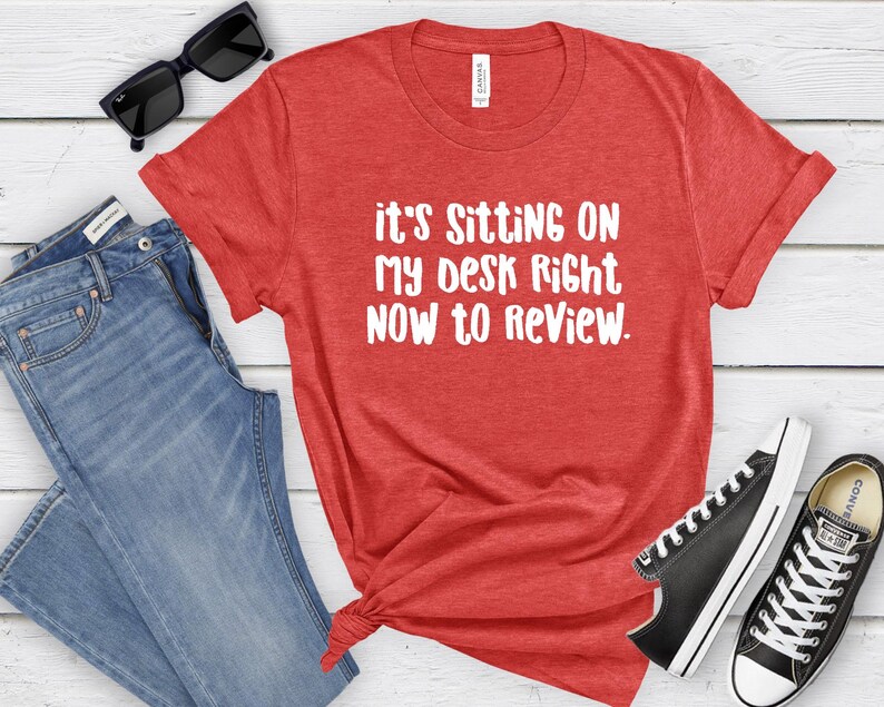 May include: Red t-shirt with the text "It's Sitting On My Desk Right Now To Review" in white. The shirt is paired with blue jeans, black sunglasses, and black Converse sneakers.