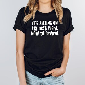 May include: Black t-shirt with white text that reads "it's SITTING ON MY DESK RIGHT NOW TO REVIEW." The shirt has short sleeves and a crew neck. The text is in a bold, sans-serif font.
