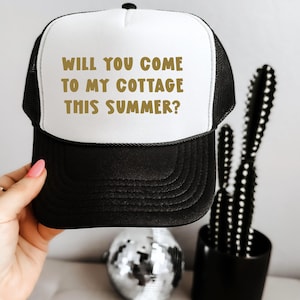 May include: Black and white trucker hat with the text "WILL YOU COME TO MY COTTAGE THIS SUMMER?" in gold lettering. The hat has a white front panel and a black brim and mesh back. A fun accessory for summer.