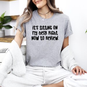 May include: A heather gray t-shirt with the text "it's SITTING ON MY DESK RIGHT NOW TO REVIEW." The shirt is a short-sleeved crew neck. The text is in a bold, black font.