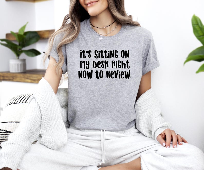 May include: A heather gray t-shirt with the text "it's SITTING ON MY DESK RIGHT NOW TO REVIEW." The shirt is a short-sleeved crew neck. The text is in a bold, black font.