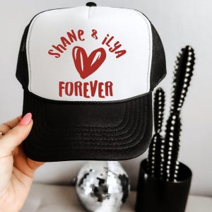 May include: A black and white trucker hat with the words "Shane & Ilya" and "Forever" in red, along with a red heart graphic. The hat has a mesh back and a curved brim.
