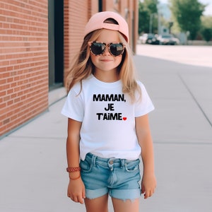 May include: A young person wearing a white t-shirt with the text "Maman, Je T'aime" in black with a red heart. The person is also wearing a pink baseball cap, sunglasses, and denim shorts.