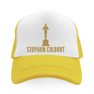 May include: Yellow and white trucker hat featuring the name "Stephen Colbert" and a gold-colored silhouette of an Oscar statuette. The hat has a mesh back and a curved brim.