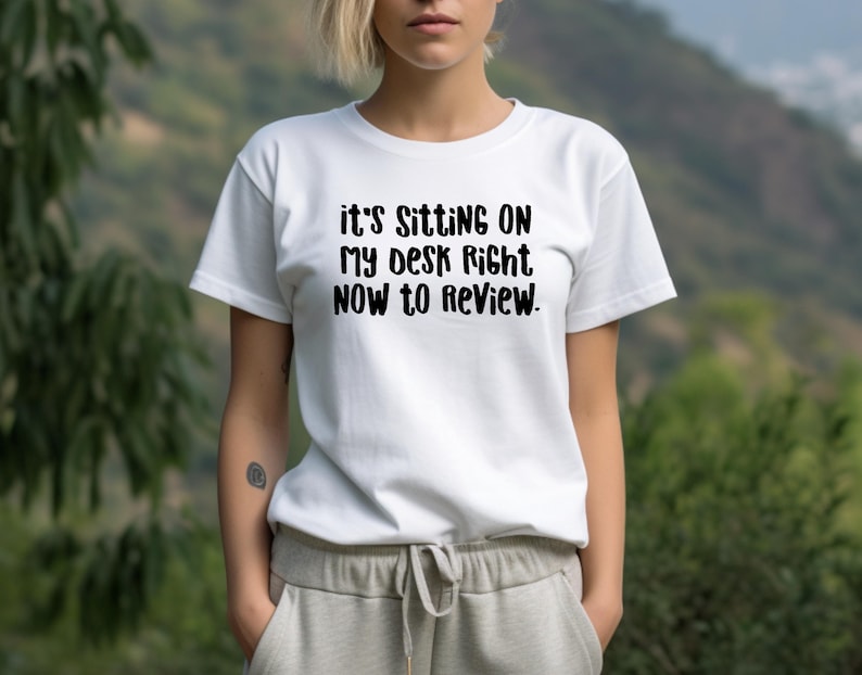 May include: White t-shirt with the black text: "it's sitting on my desk right now to review." The shirt is short-sleeved and has a crew neck. The person is wearing gray sweatpants with a drawstring.