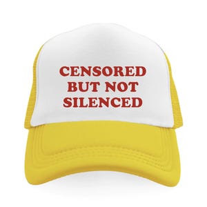 May include: A yellow and white trucker hat with the words "CENSORED BUT NOT SILENCED" printed in red block letters across the front. The hat has a mesh back and a curved brim.