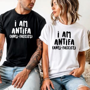May include: Black and white t-shirts with the text "I AM ANTIFA (Anti-Fascist)". The shirts are short-sleeved and worn by two people. One shirt is black, and the other is white.