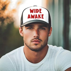 May include: A black and white trucker hat with the words "WIDE AWAKE" in red block letters. The hat has a white front panel and a black brim and mesh back. The hat is worn by a person wearing a white t-shirt.