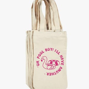 May include: A stack of beige canvas wine bottle tote bags. Each bag has a pink flamingo graphic with the text "OH. POOL BOY! I'LL HAVE ANOTHER." The bags have long handles.