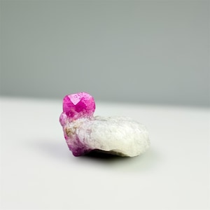 May include: A vibrant pink ruby crystal, with a faceted top, sits atop a light-colored, rough mineral base. The gemstone is the focal point, showcasing its natural beauty and color. The background is a soft, neutral gray.