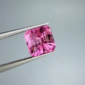 Op de afbeelding: Een levendige, vierkant geslepen roze edelsteen vastgehouden door een zilverkleurige metalen pincet. De steen heeft een gefacetteerd oppervlak dat licht reflecteert en diepe magenta en lichtere rozetinten toont in de transparante structuur.