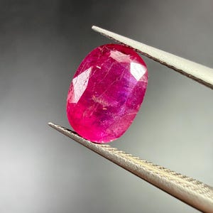以下が含まれることがあります： 金属製のピンセットで保持された、鮮やかなマゼンタレッドの楕円形カットの宝石。石の表面はファセット加工されており、光を反射し、内部のインクルージョンと半透明の構造全体にわたる深く飽和した色合いを際立たせています。