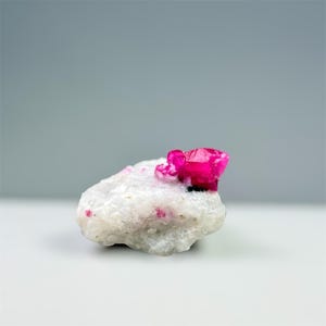 May include: A raw, natural gemstone specimen featuring a vibrant, faceted, hot pink crystal cluster atop a white, speckled matrix. The stone is set against a neutral gray background, highlighting its color and texture.