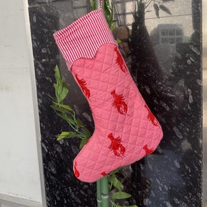 Könnte beinhalten: Rosa Weihnachtsstrumpf mit Steppmuster und rotem Hummer-Muster. Die Oberseite des Strumpfes hat rot-weiße Streifen. Eine Schlaufe zum Aufhängen ist angebracht.