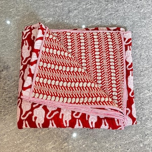 Peut inclure: Une couverture pliée à motifs rouges et blancs. La couverture présente un motif répétitif de figures d'animaux blancs sur fond rouge. La section intérieure présente un motif géométrique rouge et blanc. Les bords sont bordés d'une bordure rayée rose et blanche.