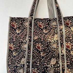 May include: A quilted tote bag with a floral pattern in black, red, and cream. The bag features long straps with a striped design and a matching trim. The bag's design is symmetrical, with the floral pattern repeating across the surface.