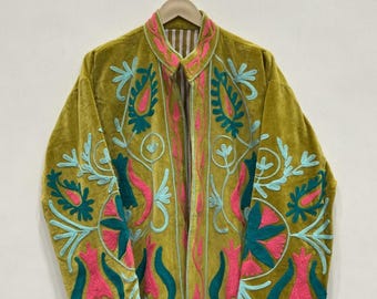 May include: Olive green velvet coat with intricate floral embroidery in teal and pink. The long coat is hanging on a wooden hanger. The embroidery design features stylized flowers and leaves, creating a bohemian aesthetic.
