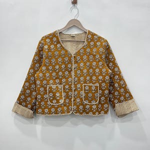 May include: A mustard yellow quilted jacket with a white floral print and a V-neck. The jacket has a white and brown striped lining and two pockets.