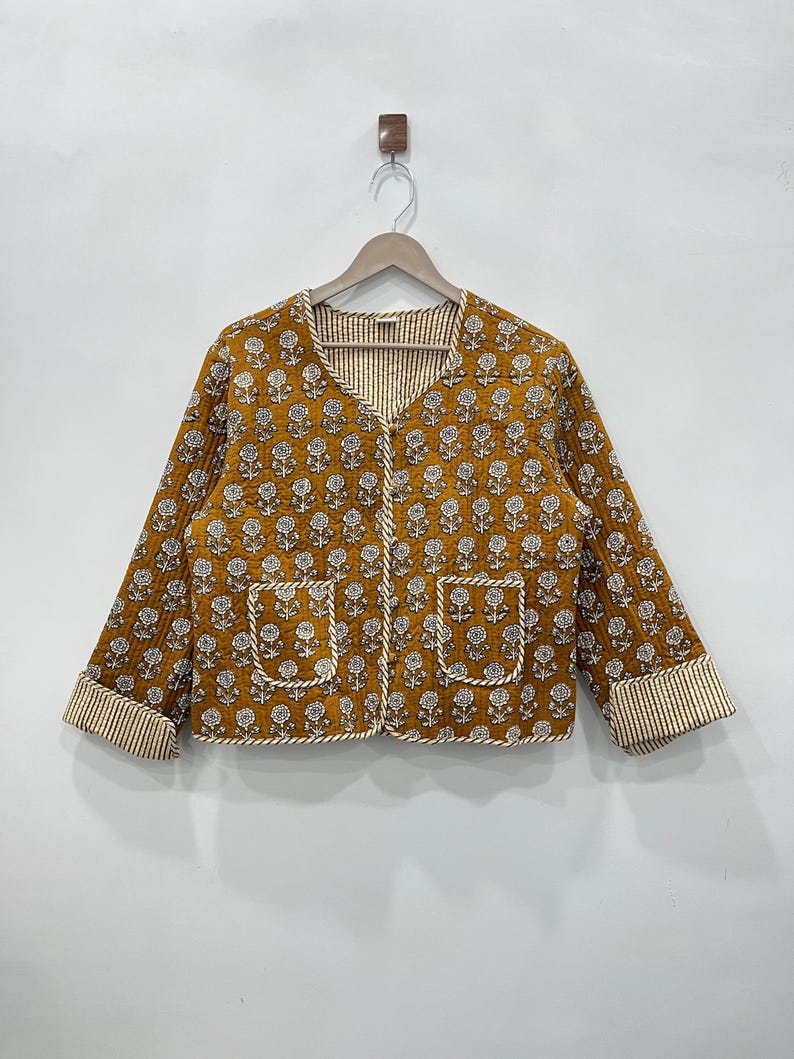 May include: A mustard yellow quilted jacket with a white floral print and a V-neck. The jacket has a white and brown striped lining and two pockets.