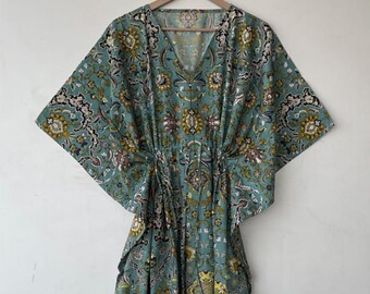 May include: A teal-colored, patterned caftan dress with a V-neck and wide sleeves. The dress features a floral and paisley design in shades of yellow, white, and brown. The waist is cinched with a drawstring. The dress is hanging on a wooden hanger.