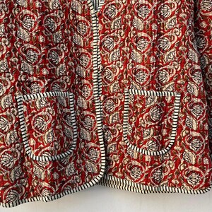 May include: A red quilted jacket with a floral print and black and white trim. The jacket has two pockets on the front.