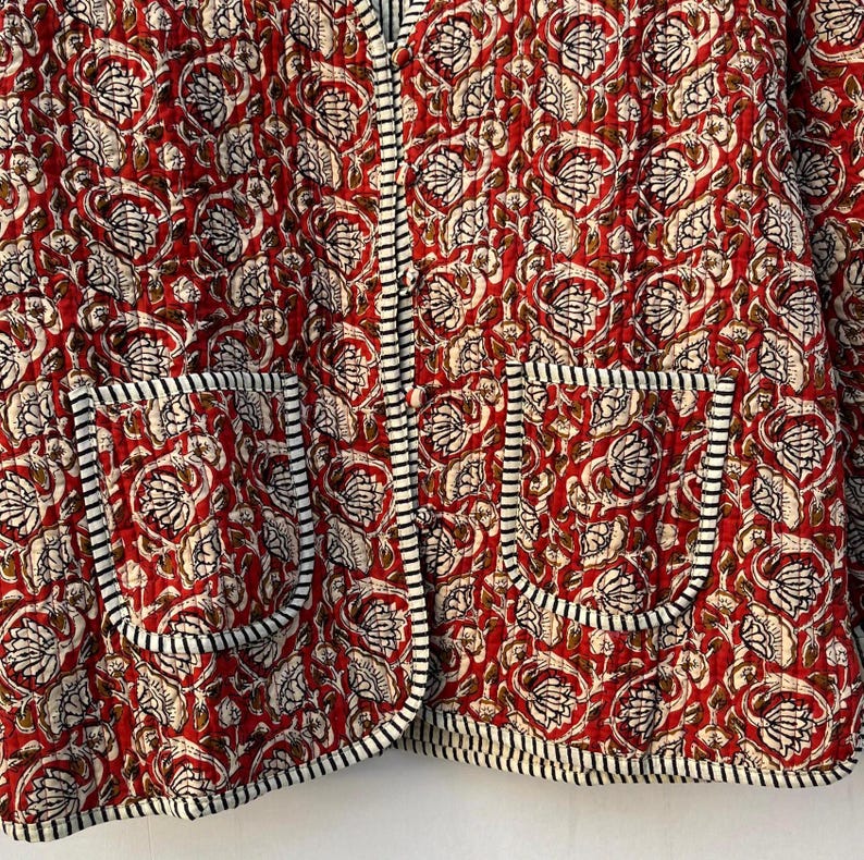 May include: A red quilted jacket with a floral print and black and white trim. The jacket has two pockets on the front.