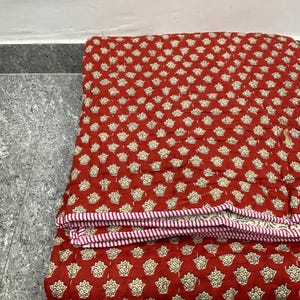 May include: A folded red quilt featuring a repeating floral design in off-white. The quilt has a striped red and white border. The quilt is folded, suggesting it is made from a soft material, and is likely for bedding or decoration.