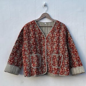May include: A red and white floral patterned quilted jacket with a V-neck and two pockets. The jacket has a button closure and a striped trim.