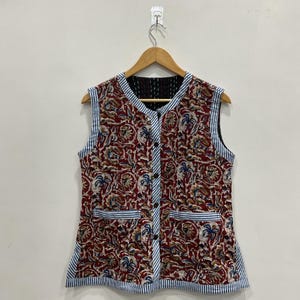 May include: Sleeveless vest with a button-up front and two pockets. The vest features a red floral pattern with blue and white accents. The edges are trimmed with blue and white striped fabric. The vest is hanging on a wooden hanger.