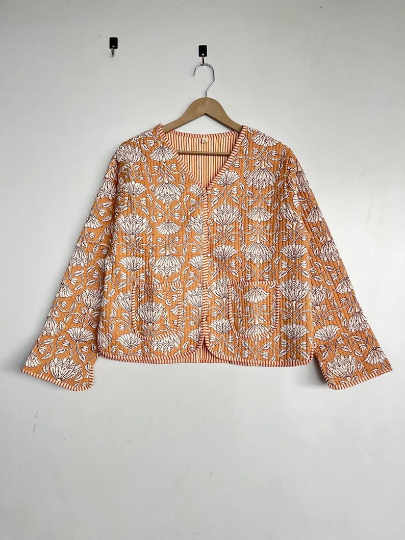 May include: A quilted jacket with a floral print in shades of orange and white. The jacket has a V-neck, long sleeves, and two pockets.