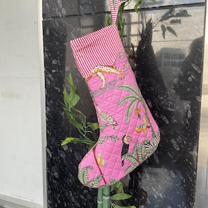 以下が含まれることがあります： ピンク色のクリスマスストッキング。ヒョウ、オオハシ、シマウマをあしらったキルティングデザイン。ストッキングの上部は赤と白のストライプ。吊り下げ用の緑と白のストライプのループが付いています。