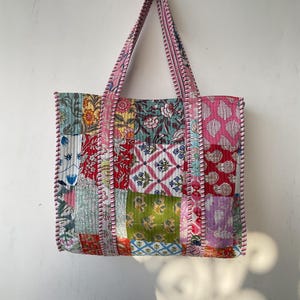 May include: A patchwork tote bag with a pink and white striped handle. The bag is made of different fabrics with floral and geometric patterns in shades of pink, red, green, blue, and yellow.