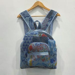 May include: A blue patchwork backpack with floral and geometric patterns. The backpack has a small front pocket and adjustable straps. It is hanging on a wooden hanger against a white wall.