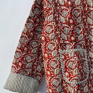 May include: A red quilted jacket with a floral print and black and white striped trim. The jacket has a pocket on the left side and a V-neck.