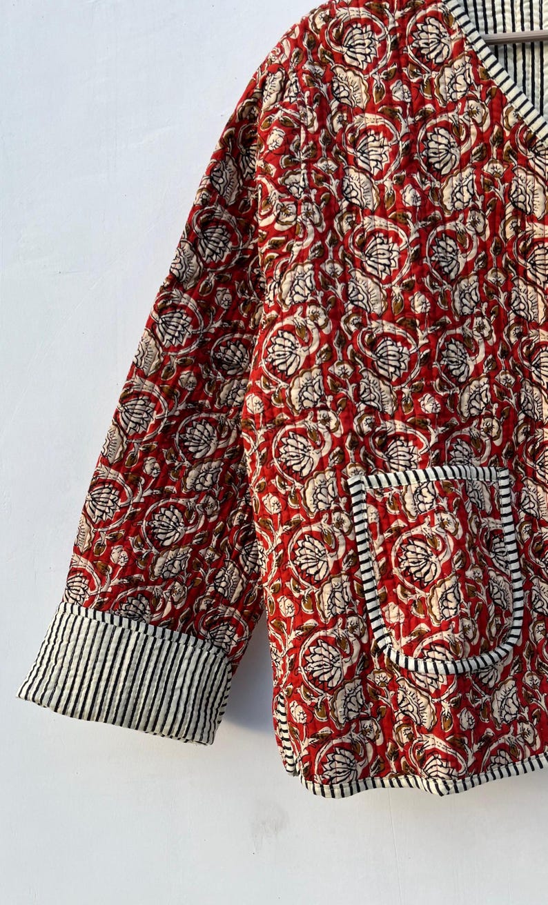 May include: A red quilted jacket with a floral print and black and white striped trim. The jacket has a pocket on the left side and a V-neck.