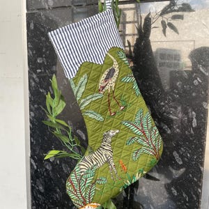 Puede incluir: Un calcetín navideño acolchado con un fondo verde que presenta un diseño de cebra y pájaro. La parte superior del calcetín tiene un patrón de rayas blancas y azul marino. El calcetín está decorado con un tema de la jungla.