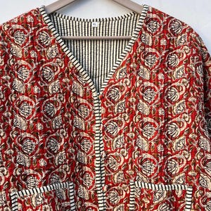 May include: A red quilted jacket with a floral print and black and white striped trim. The jacket has a V-neck and two pockets. The size tag says "XL".