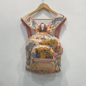 Può includere: Zaino colorato con un design floreale e fantasia. Lo zaino ha una tasca frontale e spallacci regolabili. I colori principali sono crema, blu, giallo e rosso. Lo zaino è appeso a una gruccia di legno.