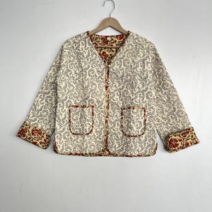 May include: A cream-colored quilted jacket with a floral print. The jacket has a V-neck and two pockets. The cuffs and collar are lined with a red and yellow floral print.