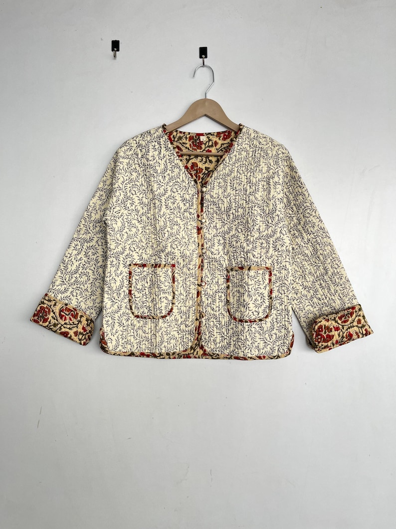 May include: A cream-colored quilted jacket with a floral print. The jacket has a V-neck and two pockets. The cuffs and collar are lined with a red and yellow floral print.