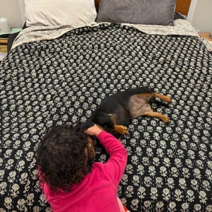 May include: A black and white floral patterned quilt covers a bed. A black dog is lying on the quilt. A person wearing a pink shirt is interacting with the dog. Pillows are visible at the head of the bed.