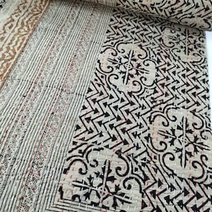 以下が含まれることがあります： 幾何学模様のベージュとブラックのカンタキルト。キルトは半分に折り畳まれ、複雑なステッチのクローズアップが示されています。