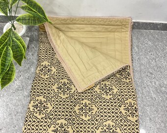May include: A rectangular quilted blanket featuring a tan base with a repeating black geometric and floral pattern. The blanket is folded at the top corner to reveal a solid tan underside with a stitched grid design and a thin striped border.