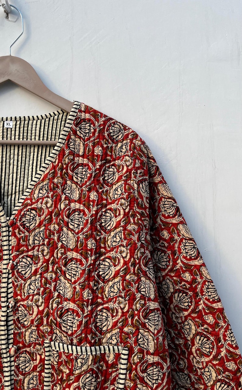 May include: A red and white floral quilted jacket with a black and white striped trim. The jacket has a V-neck and a button closure. The size tag says "XL".