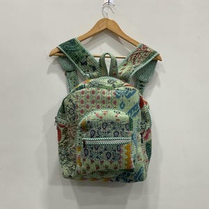 Op de afbeelding: Een lichtgroene rugzak met een patchwork-ontwerp. De rugzak heeft een voorvak met een ritssluiting en verstelbare bandjes. De stof heeft een verscheidenheid aan kleurrijke patronen, waaronder bloemen en geometrische vormen. Hij hangt aan een houten hanger.