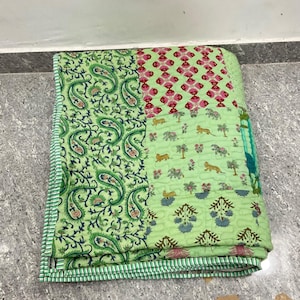 May include: A folded quilt with a patchwork design. The quilt features sections with paisley, floral, and animal prints in shades of green, pink, and blue. The edges are finished with a striped border.