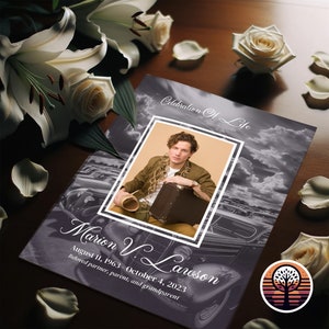 May include: A memorial card with a photo of a person, the text "Celebration Of Life", and the name "Marion V. Lawson" with dates. White roses and lily flowers are arranged around the card on a wooden surface.