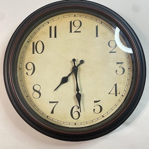Peut inclure: Une horloge murale ronde avec un cadre marron foncé et un cadran crème. L'horloge présente des chiffres romains noirs et des aiguilles noires, indiquant qu'il est environ 7h30. L'horloge a un design classique et vintage.