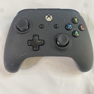 May include: A black Xbox controller featuring a directional pad, two analogue sticks, and coloured buttons. The controller has the Xbox logo and several small buttons. The controller is on a white surface.